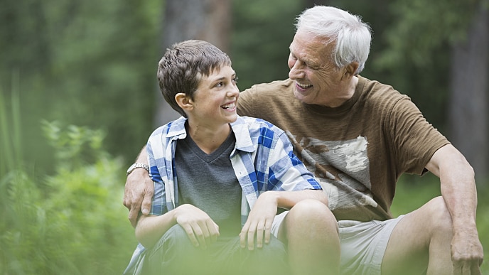 um homem com idade avançada, cabelos grisalhos, vestindo camiseta manga curta na cor marrom, sorri enquanto abraça um menino pré-adolescente que também sorri, tem cabelos curtos e escuros, vestindo uma camiseta por baixo de uma camisa xadrez