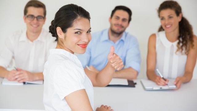 Mulher confiante dando um sinal de positivo durante uma entrevista de emprego, enquanto os entrevistadores observam sorrindo ao fundo.