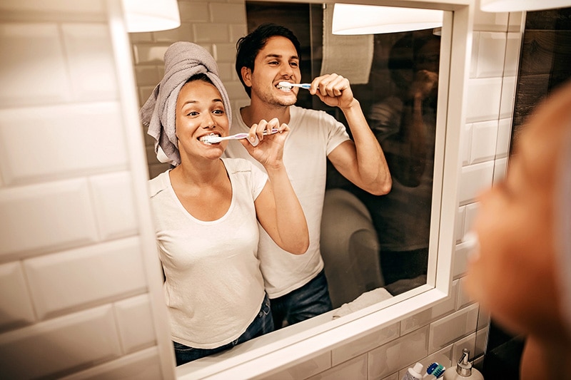 Casal feliz em frente ao espelho escovando os dentes com uma pasta de dente sem glúten