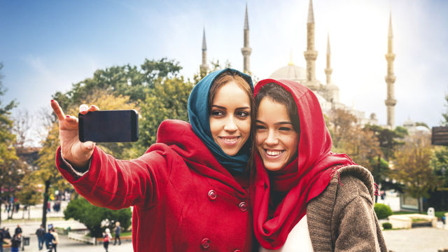 duas mulheres tirando uma selfie. A mulher à esquerda veste casaco vermelho e tem cabelos claros. A mulher à direita veste um casaco marrom com uma blusinha branca por baixo. Ambas estão sorrindo.
