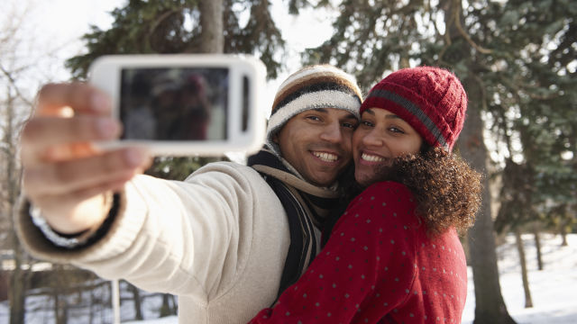 primeira imagem: casal se abraçando e sorrindo para a câmera do celular, enquanto tiram uma selfie. A mulher está vestindo um casaco para frio intenso e gorro, ambos na cor vermelha. O homem está vestindo casaco para frio intenso na cor creme com detalhes marrons, o gorro dele é marrom com branco.