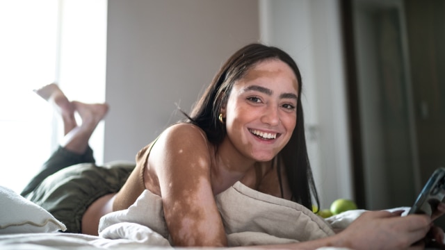 Mulher jovem deitada na cama sorrindo enquanto segura o celular nas mãos