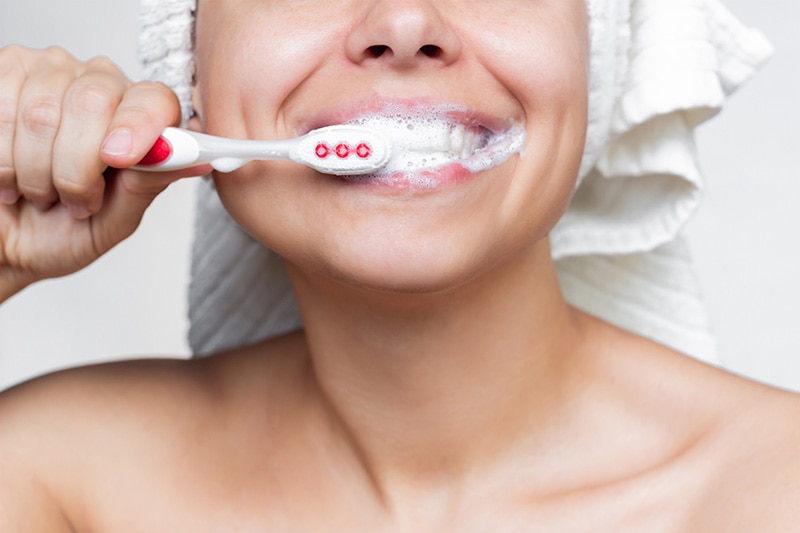 Mulher com toalha de banho na cabeça escovando os dentes, feliz porque sabe que tem proteção total com Colgate Total 12