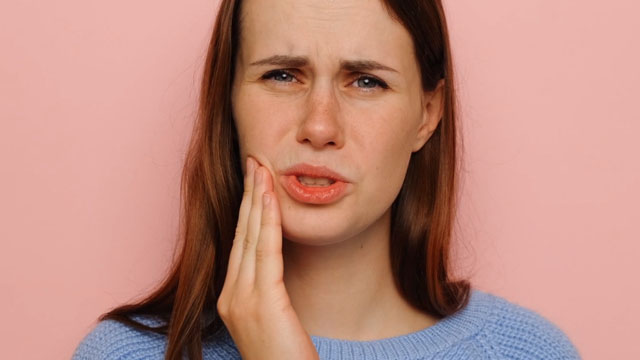 Mulher com a mão na bochecha e com a cara de dor por causa de uma lesão bucal